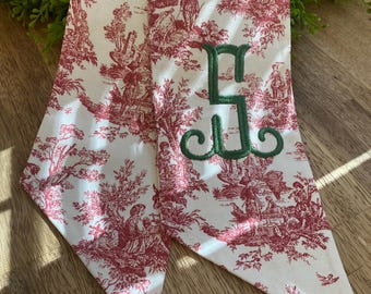 May include: A white and red toile scarf with a green embroidered letter "S". The scarf features a classic pastoral scene design. The scarf is made of a soft fabric and is a stylish accessory.