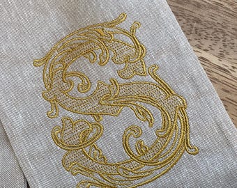 May include: Close-up of a linen table runner with an embroidered gold letter "S". The ornate script is centered on the light beige fabric. The runner is partially visible, with a wooden surface in the background.