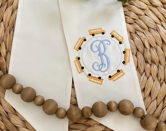 May include: Cream-colored fabric banner with a light blue embroidered letter "B" surrounded by yellow school bus designs. A wooden bead garland and greenery are also visible.