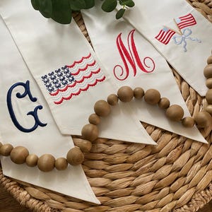 May include: Three off-white fabric banners featuring embroidered designs. One banner displays a navy blue letter G and a wavy American flag. The second banner features a red letter M. The third banner shows two small American flags tied with a light blue bow.