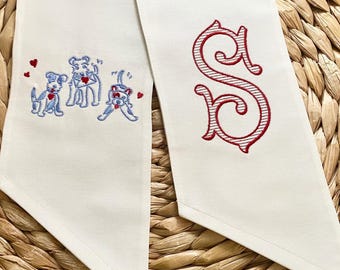 May include: Two white fabric ribbon ties with embroidered designs. One tie features three blue dogs with red hearts and the other tie features a red embroidered letter "S".