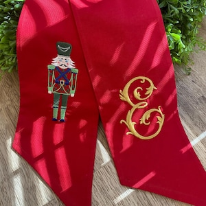 Puede incluir: Una bufanda roja brillante con un soldado cascanueces bordado y una letra "E" dorada. El cascanueces está detallado con acentos verdes, rojos y azules. La bufanda está hecha de un material liso, posiblemente satinado.