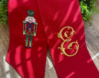 May include: A bright red scarf with an embroidered nutcracker soldier on one end and a gold letter "E" on the other. The nutcracker is detailed with green, red, and blue accents. The scarf is made of a smooth, possibly satin-like material.