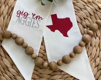 May include: White fabric graduation sash with the text "gig 'em Aggies" embroidered in gray and a red outline of the state of Texas.