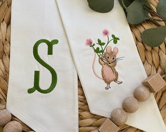 May include: Two white fabric panels with green embroidery. One panel has the letter "S" embroidered in green. The other panel has a small mouse holding a pink clover embroidered in green. The panels are on a woven wicker surface with a string of wooden beads.