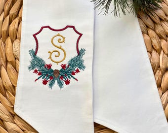 May include: White fabric place card holder with a gold embroidered letter "S" in a red and gold shield surrounded by green pine branches and red berries.