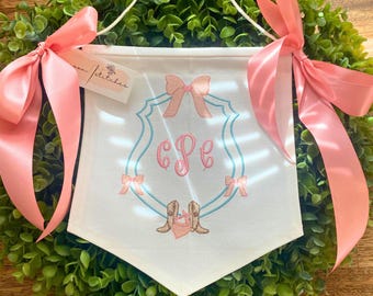 May include: A white banner with a pink and blue embroidered crest featuring the initials "CPE" and a bow. The banner is decorated with pink ribbons and hangs from a wreath of green foliage.