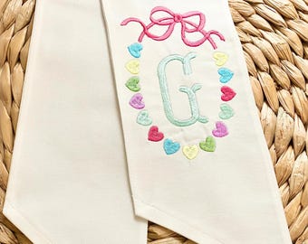 May include: White fabric sash with a pink bow and a green letter G surrounded by a circle of colorful hearts.