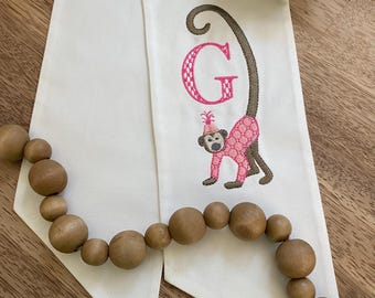 May include: White fabric table runner with a pink embroidered letter "G" and a monkey wearing a party hat. A wooden bead garland is draped across the runner. The monkey is wearing a pink outfit.