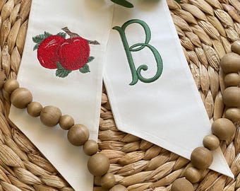May include: Two white fabric table runners with embroidered designs. One features two red apples with green leaves, the other a green letter "B". A wooden bead garland and greenery are also visible.