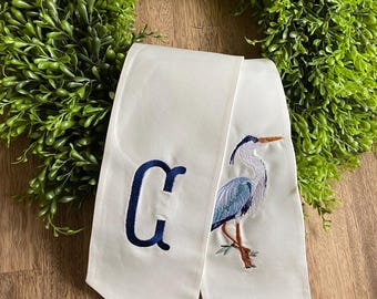 May include: A white fabric sash featuring an embroidered navy blue letter G on the left side and a detailed, multi-colored heron bird illustration on the right side. The sash has pointed ends and is displayed against a green leafy wreath.