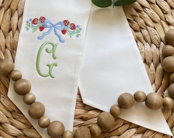 May include: White fabric ribbon with a green embroidered letter "G" and a blue bow with red embroidered strawberries. The ribbon is on a woven wicker surface.