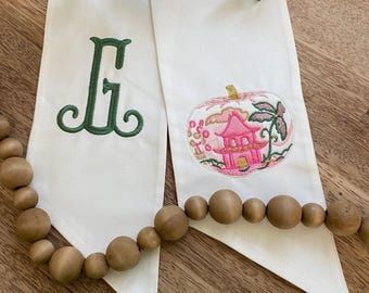 May include: Two white fabric ribbons with embroidered designs. One has a green letter "G", the other a pink pagoda design. A wooden bead garland and greenery are also visible.