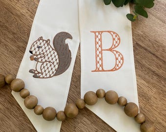 May include: Two off-white fabric banners with embroidered designs. One features a squirrel holding a nut, the other a letter "B" in orange. A wooden bead garland and green foliage are also present.