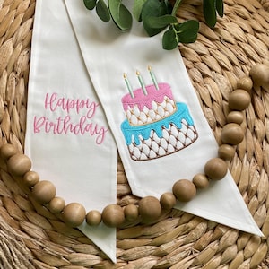 May include: White fabric banner with pink and blue embroidered cake with candles and the text "Happy Birthday".