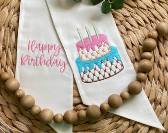 May include: Two white fabric pennants featuring embroidery. One pennant displays the pink stitched text Happy Birthday. The other pennant features an embroidered two-tier cake with pink and blue icing, a brown lattice pattern, and three green candles.