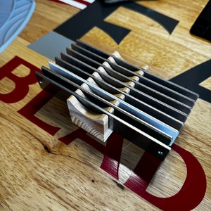 Może przedstawiać: Drewniany i metalowy stojak na ostrza do golenia z ośmioma szczelinami do przechowywania ostrzy. Stojak stoi na drewnianej powierzchni z napisem "BLADE" nadrukowanym na czerwono.