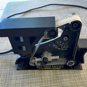 Könnte beinhalten: Schwarzer Bandschleiferaufsatz mit einem Holzband, einem Spannknopf und einem Zifferblatt mit Markierungen von 10 bis 35. Der Aufsatz ist mit "Ben Onion Edition Blade Grinding Attachment" beschriftet.