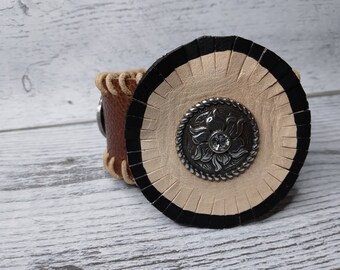May include: A brown leather cuff bracelet with a white leather circle and black trim. The circle has a silver metal flower design with a rhinestone in the center.