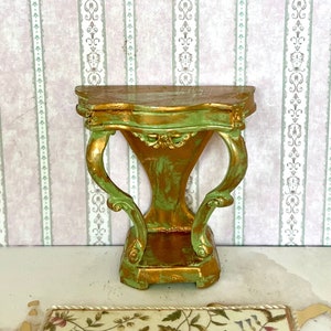 May include: A miniature green and gold console table with a gold ornate frame with a portrait of a woman in a pink dress. The table is on a white and gold patterned rug.