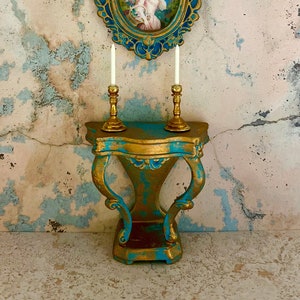 May include: A teal and gold console table with two candlesticks and a framed portrait of a young person in a white dress. The wall behind the table is textured and painted in shades of pink and blue.