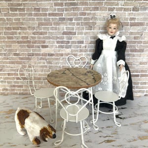 May include: Miniature dollhouse furniture set with a round table, three chairs, and a porcelain doll. The table has a marble-like top. The chairs are white with heart-shaped backs. A small dog figurine is also present.
