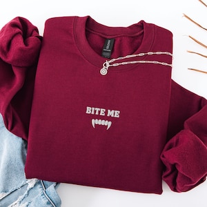 May include: A burgundy sweatshirt with the text "BITE ME" and fangs embroidered in white. A silver necklace with a pendant is draped across the sweatshirt. The sweatshirt is paired with blue jeans.