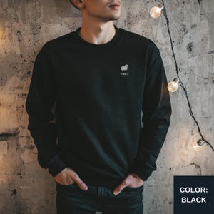 May include: Black crew neck sweatshirt with a small white sheep graphic and the text "Hola Z)". The sweatshirt is a solid black color and has long sleeves. The color black is displayed in a black box in the bottom right corner.