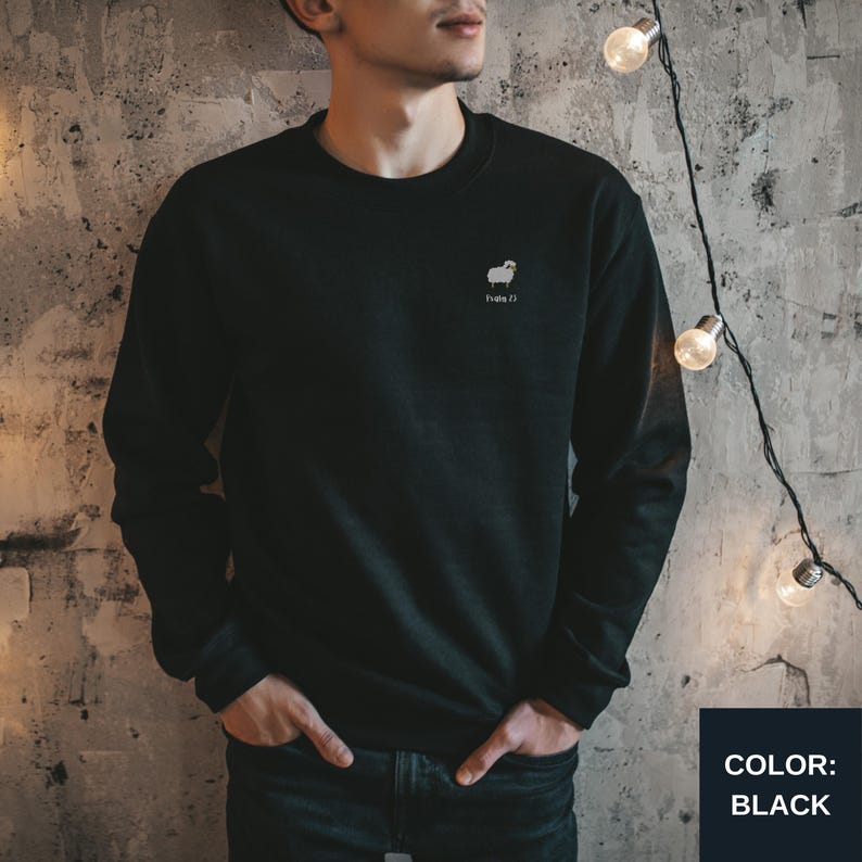 May include: Black crew neck sweatshirt with a small white sheep graphic and the text "Hola Z)". The sweatshirt is a solid black color and has long sleeves. The color black is displayed in a black box in the bottom right corner.
