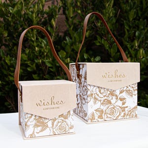 Peut inclure: Deux boîtes cadeaux beiges avec des poignées en cuir marron. Les deux boîtes ont un fond blanc avec un motif floral doré et les mots "wishes a gift for you" imprimés en or.