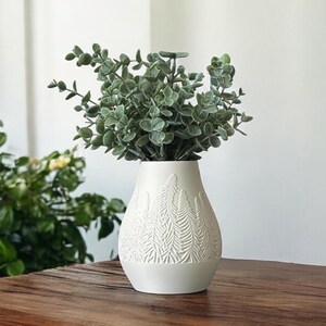 May include: White ceramic vase with a textured, fern-like pattern. The vase is filled with a bouquet of green eucalyptus leaves.