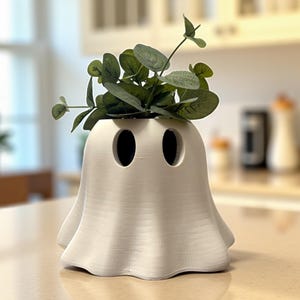 May include: A white ghost-shaped planter with two eye-shaped openings and a ruffled bottom. The planter is filled with green leafy sprigs. The planter is sitting on a light-colored countertop.