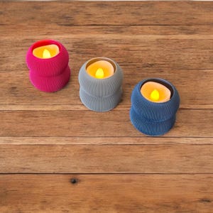 May include: Three decorative candle holders with a ribbed design. The candle holders are in shades of pink, gray, and blue. Each holder has a small, round, yellow LED candle inside.