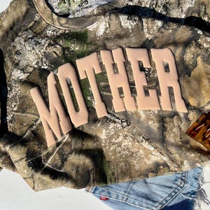 May include: A camouflage long-sleeve shirt with the word "MOTHER" in tan letters. A denim skirt and a tortoiseshell hair clip are also visible. The shirt has a green and brown pattern.