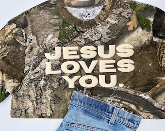 May include: A cropped camouflage t-shirt with the text "JESUS LOVES YOU" in large, off-white letters. The shirt is paired with a section of blue denim jeans. The shirt has a Realtree logo on the collar.