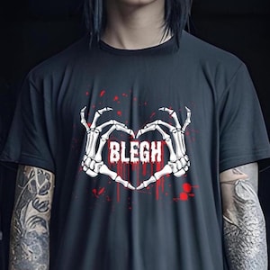 以下が含まれることがあります： 黒いTシャツに、2つの骸骨の手がハートの形を作り、その中に「BLEGH」という文字が入ったグラフィックがプリントされています。グラフィックは赤と白です。