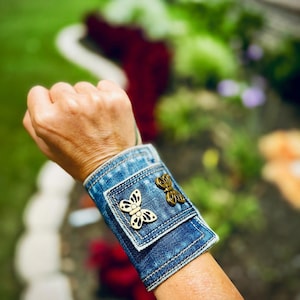 Puede incluir: Un brazalete de denim con dos abalorios de mariposa dorados. El brazalete está hecho de denim azul y tiene un borde sin rematar.
