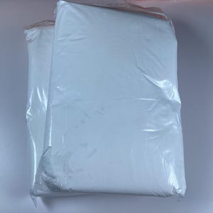 May include: Two large, rectangular, white packages sealed in clear plastic. The packages are stacked next to each other. The plastic is slightly wrinkled, and the contents are not visible. The packages are likely filled with a soft material.