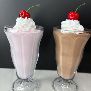 以下が含まれることがあります： ピンクと茶色の2つのミルクシェイクグラス。ホイップクリームと赤いさくらんぼがトッピングされています。透明なグラスはフレア型で、台座の上に置かれています。ダイナーやレトロなテーマに最適です。