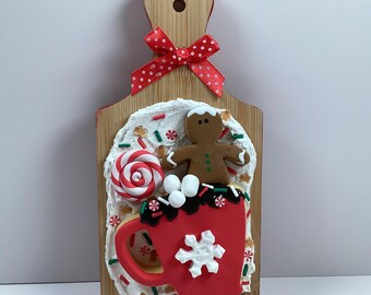 May include: A wooden cutting board with a red bow and a decorative Christmas scene. The scene includes a red mug with a snowflake, a gingerbread man, a lollipop, and sprinkles.