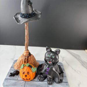 May include: A Halloween-themed figurine set featuring a witch's hat with a spider, a broom, a carved orange pumpkin, a black cat, and a small black rat. The figurines are arranged on a gray platform.