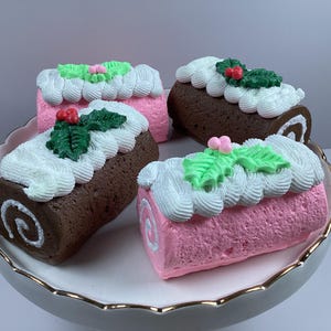 May include: Four decorative Yule log cakes on a white and gold-rimmed serving plate. Two are chocolate brown, and two are pink. Each is topped with white frosting, green holly leaves, and red berries. The cakes are a festive holiday decoration.