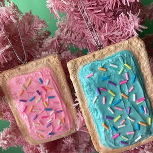 May include: Two frosted pop-tart ornaments with colorful sprinkles. The pop-tart on the left has pink frosting and the pop-tart on the right has blue frosting.