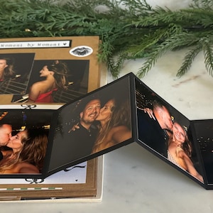 May include: A scrapbook with photos of a couple, including a multi-panel photo display. The scrapbook has a kraft paper cover with black spiral binding. The photos show the couple kissing and smiling.