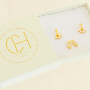 May include: A white box with a gold logo that says "Charm Her" and two pairs of gold earrings with clear crystals. One pair is a flower design and the other is a teardrop design.