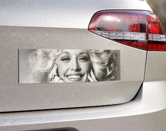 May include: A rectangular bumper sticker featuring a black and white photographic portrait of a smiling person with voluminous blonde hair, hands positioned near the face, and rings on the fingers, applied to the silver trunk of a vehicle.