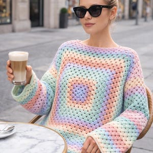 May include: A pastel-coloured, square-patterned crochet sweater. The sweater features a palette of soft colours including pink, blue, yellow, and green. The person is wearing black sunglasses and holding a glass of latte.