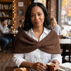 May include: A woman wearing a brown knitted shawl over a white shirt, seated at a cafe table. She holds a cup of coffee, with a small plate of cake nearby. The background includes a bookshelf and a window.