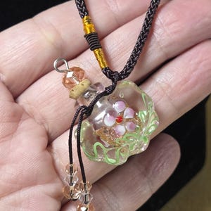 May include: A clear glass pendant necklace with a floral design. The pendant features pink and red flowers with gold accents and green leaves. The necklace has a brown cord with gold and brown accents and clear and peach-colored beads.