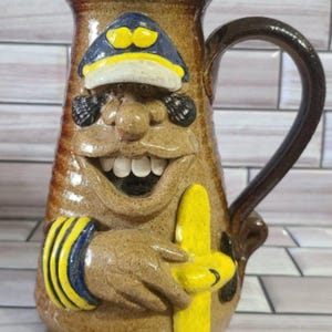 May include: A brown ceramic pitcher featuring a sculpted face with a wide, toothy grin, wearing a blue and yellow captain's hat. The figure has a hand with yellow and blue striped cuffs holding a yellow object, with a dark brown handle on the side.